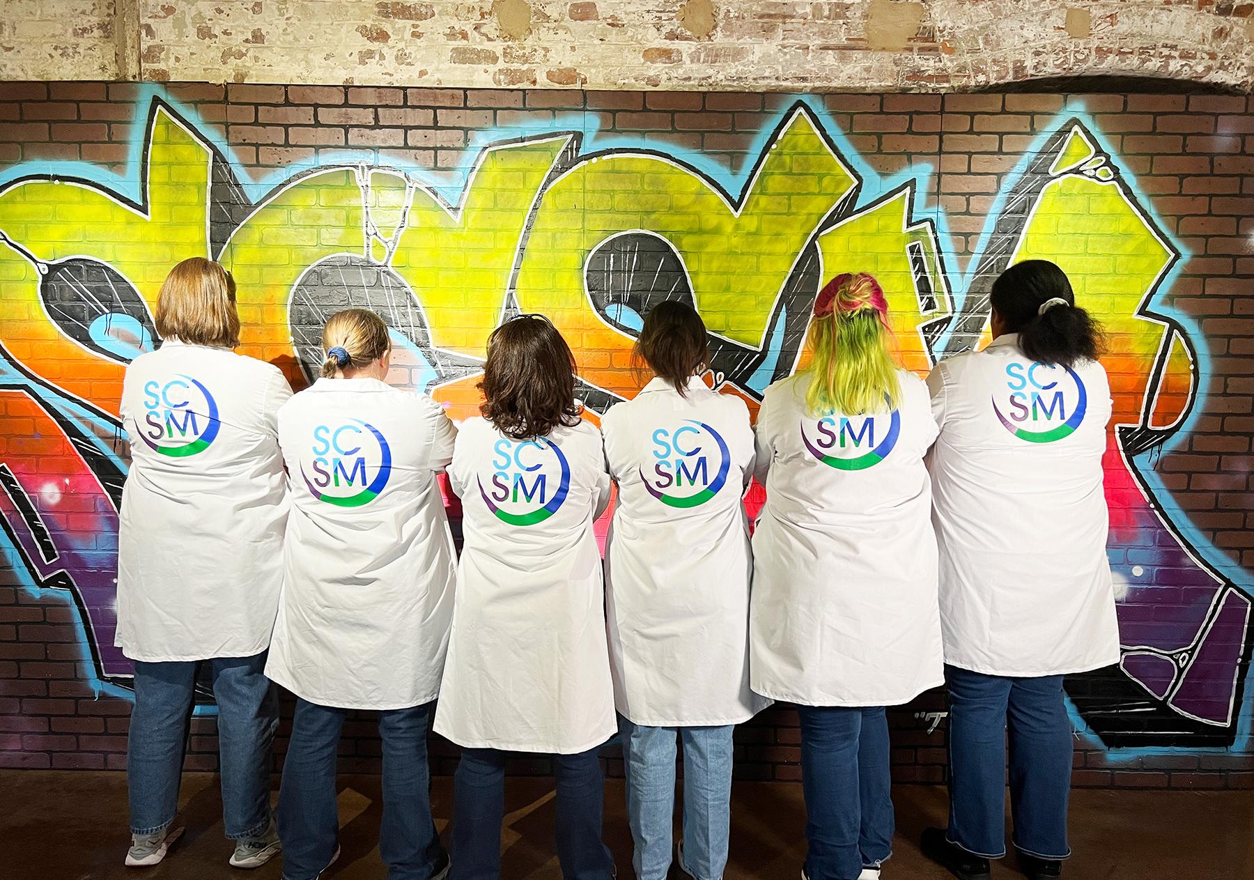 Group stand with backs to camera showing the SCSM logo on the backs of their lab coats