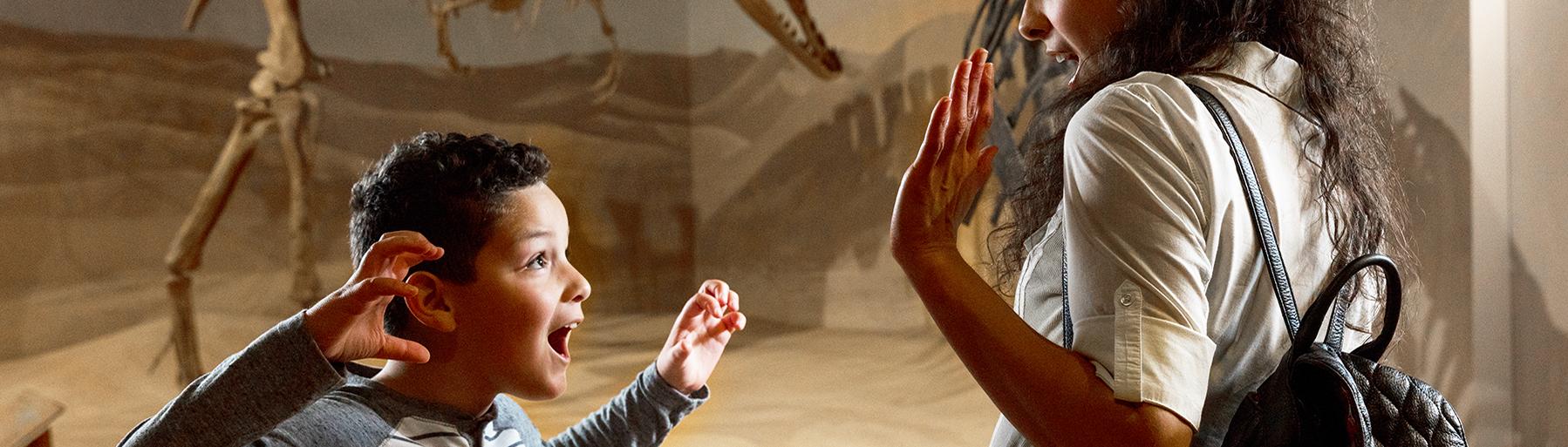 Woman and young boy stand in front of dinosaur skeleton as the boy makes a 'Rawr' face with hands up like dinosaur claws in the direction of the woman
