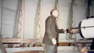Man in brown suit jacket stands with hand on the eye piece of a large white telescope