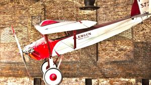 White biplane with red trim and nose that reads 'Clemson' on the side hangs from ceiling on display at the State Museum