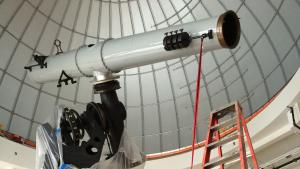 Man facing away from camera stands holding a rope attached to the cylinder of a large white telescope above him