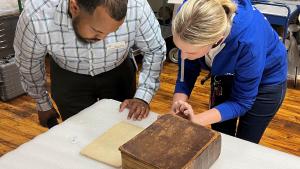 A man and woman bend down towards a table to examine a large, old leather bound book