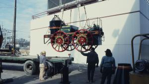 Life size antique train engine car is suspended on a lift as it is moved from the back of a flatbed truck to the ground. 