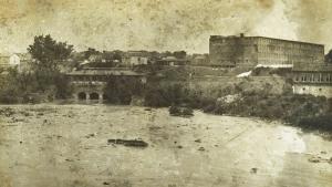 Old, sepia toned photograph shows canal in the foreground with large brick building on a rise above the canal