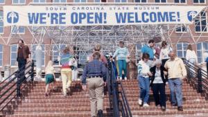 Group of people walking up stairs towards a large brick building with a banner over the entrance that reads "We're Open! Welcome!"