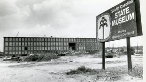 Black and white image of long, four story textile mill building under renovation. A sign is posted in front that reads "South Carolina State Museum Opening Windows to Our World".