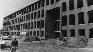 Black and white image of long, four story textile mill building under renovation. An man in a hard hat is visible in the lower left corner.