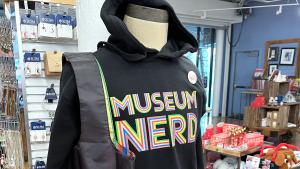 Display Mannequin Wearing a Black hooded Sweatshirt and Black tote bag that both read in multi colored lettering Museum Nerd