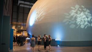 Wide shot of the planetarium lobby showing a group of reception guests dressed in cocktail attire talking with the planetarium just behind.