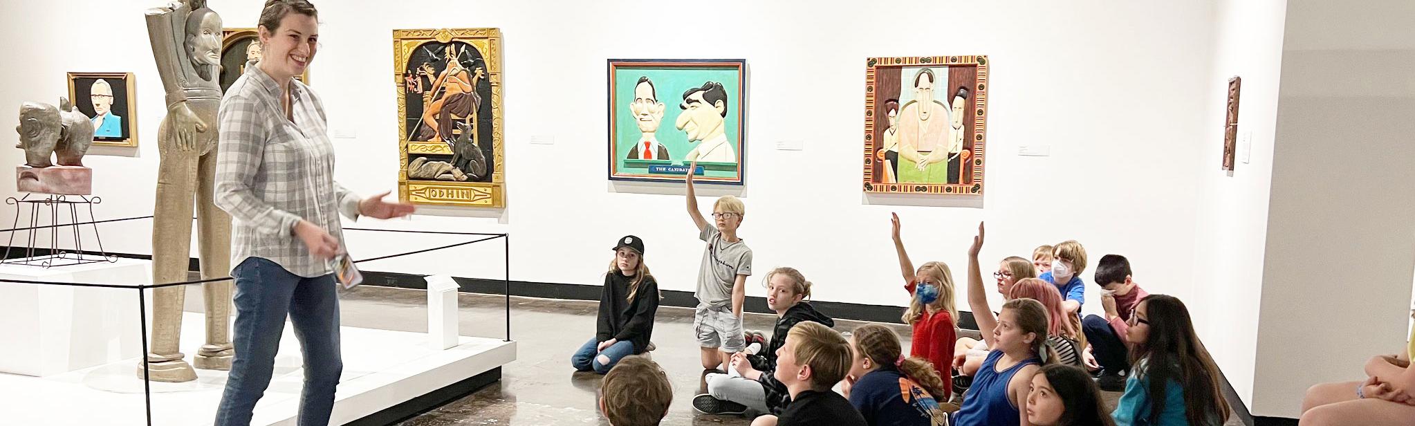 Curator of Art stands in from of a group of children, some raising their hands, seated on the floor in the Lipscomb Art Gallery. 