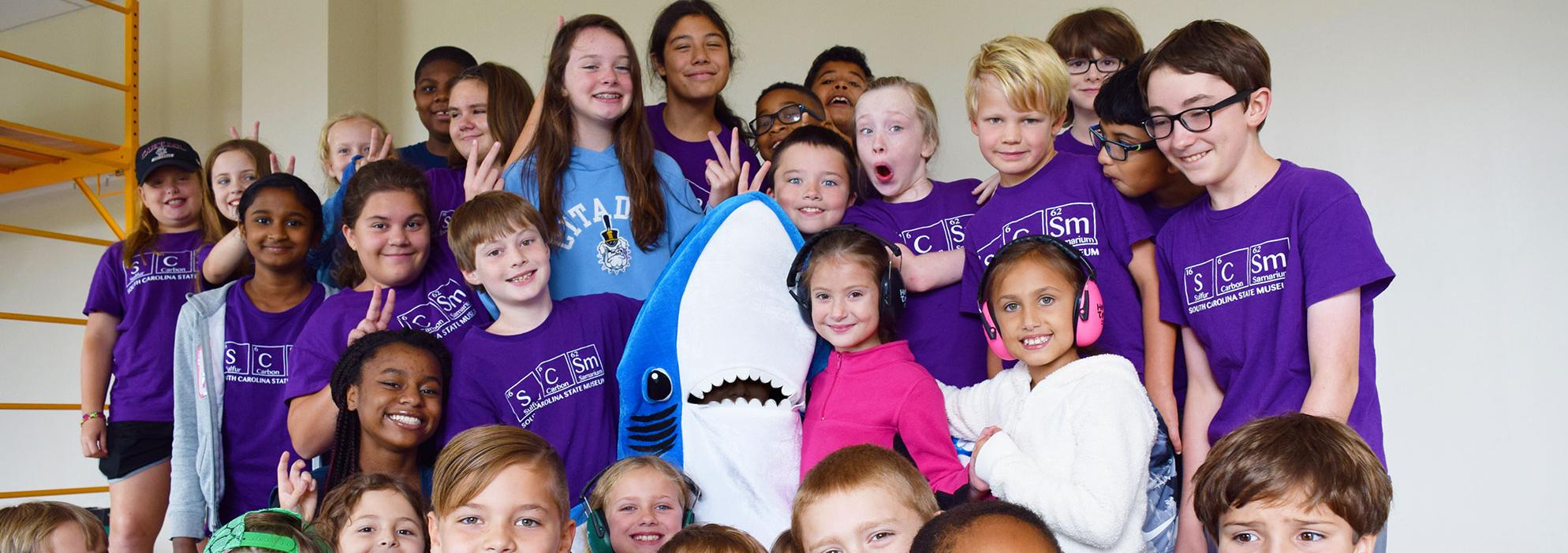 Large group of kids aged 6-12 wearing purple SCSM camp shirts posing with an educator in a shark mascot costume.
