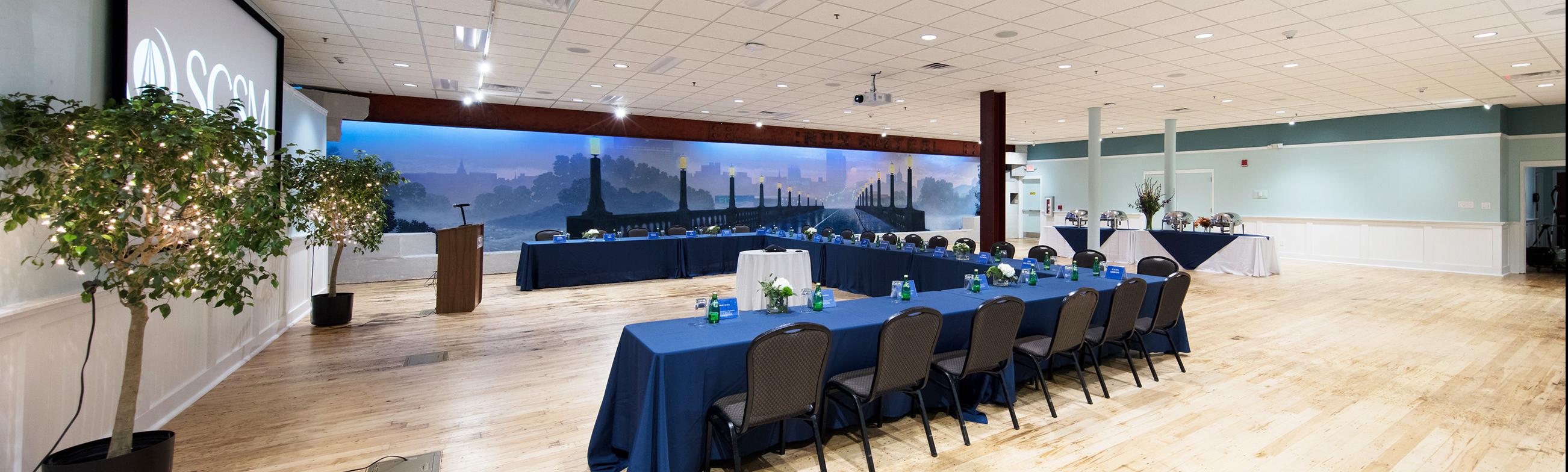 Large room with light colored wooden floor has table set up in u shape facing a large screen on the wall with mural of bridge in the background