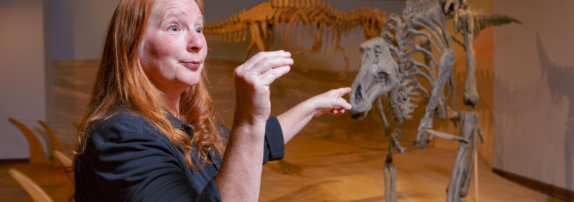 Woman stands in front of display of replica life-size dinosaur fossil skeletons with her arm and hand making a upside down L shape
