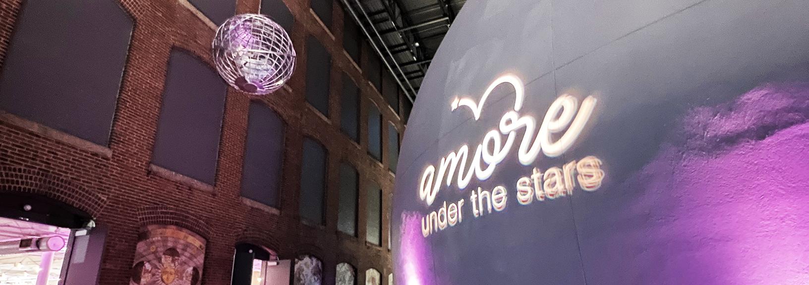 Exterior of planetarium dome and lobby with the Amore Under the Stars event logo projected in white lights against the gray dome with pink up lights to either side of the logo