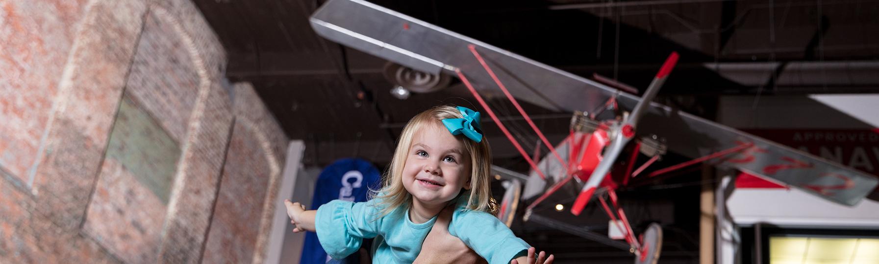 Father holds small daughter aloft as she spreads her arms like wings with the Clemson Airplane visible suspended from the ceiling behind her