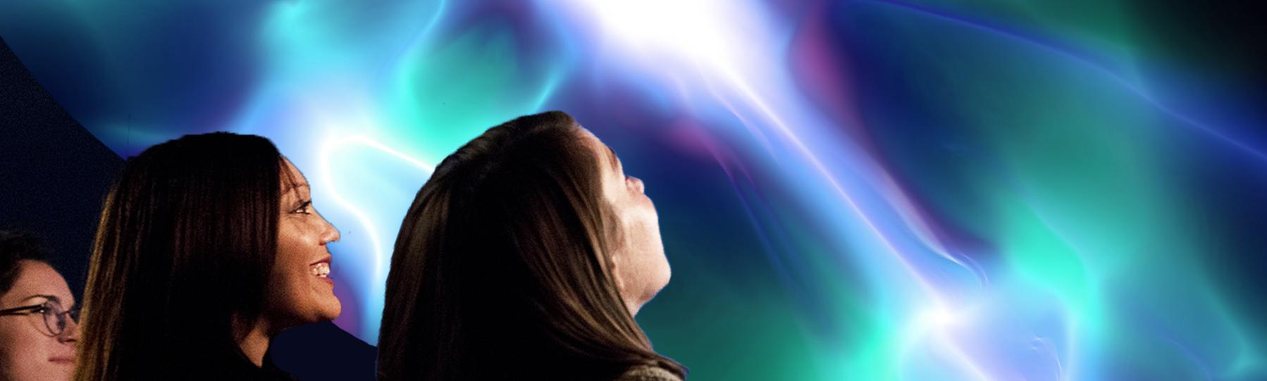 Close up of the profiles of 3 women as the look up at the planetarium dome which displays colorful lights and lasers.