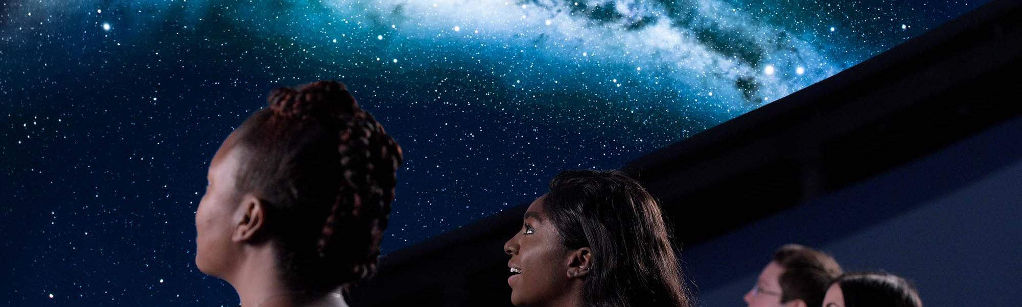Close up image of four people shown from a back/side profile as they look up to watch a projection of the Milky Way in a planetarium dome