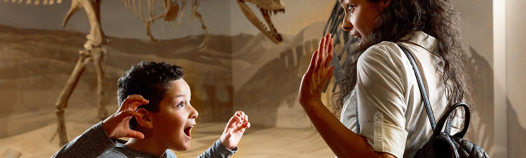 Woman and young boy stand in front of dinosaur skeleton as the boy makes a 'Rawr' face with hands up like dinosaur claws in the direction of the woman