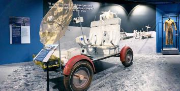 Open vehicle with two large white seats and gold mesh satellite attached to front and four red capped wheels sits on display with a replica of the moon's surface beneath it.