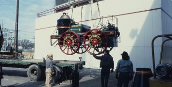 Life size antique train engine car is suspended on a lift as it is moved from the back of a flatbed truck to the ground. 