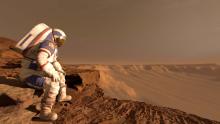 Astronaut sits wearing a white spacesuit with a red, dusty landscape visible behind them