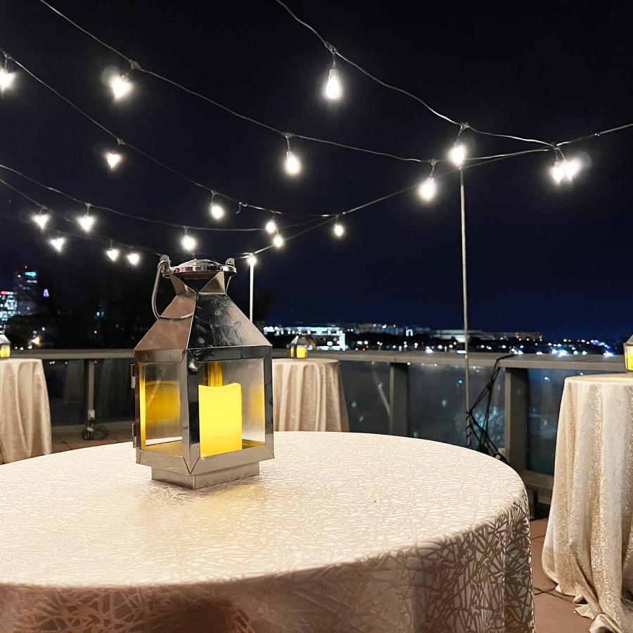 Terrace at night with skyline visible in background and string lights set up above three formally set tables