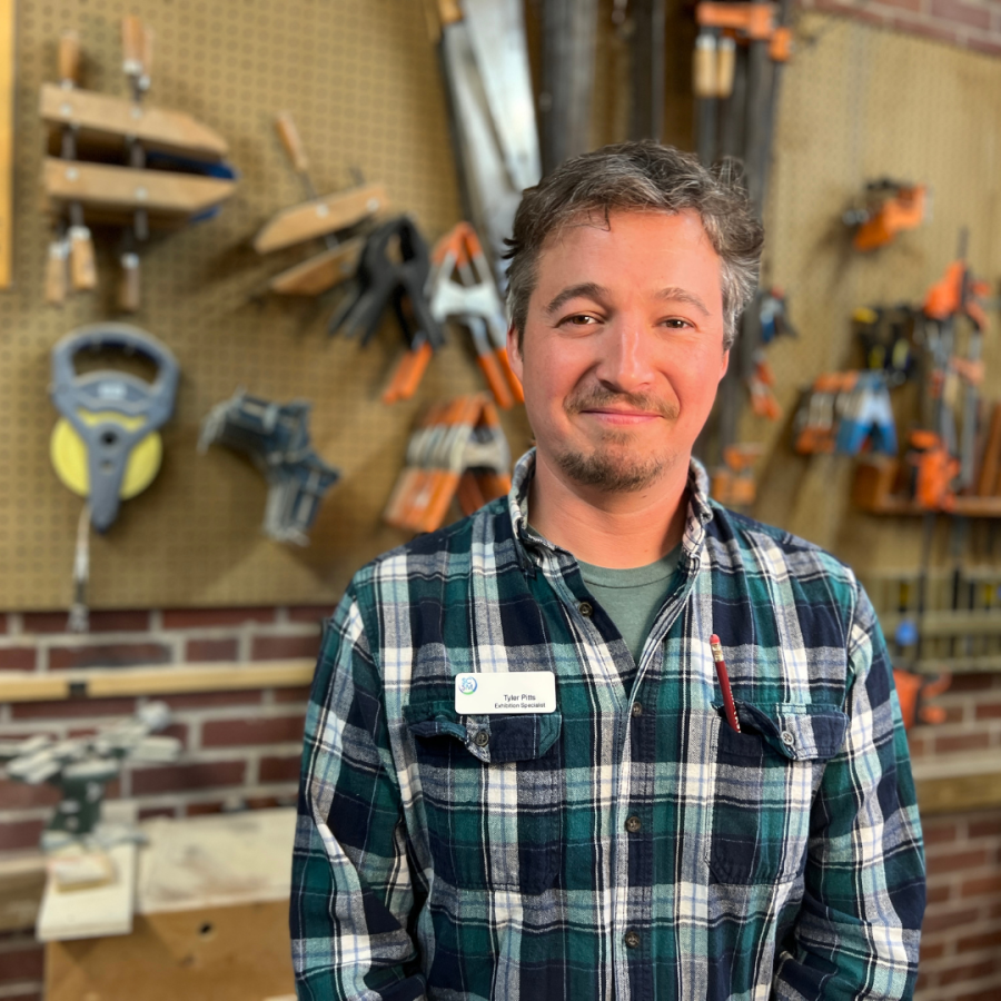 Man with beard and mustache wearing green plaid shirt smiles at camera with wall of tools behind him