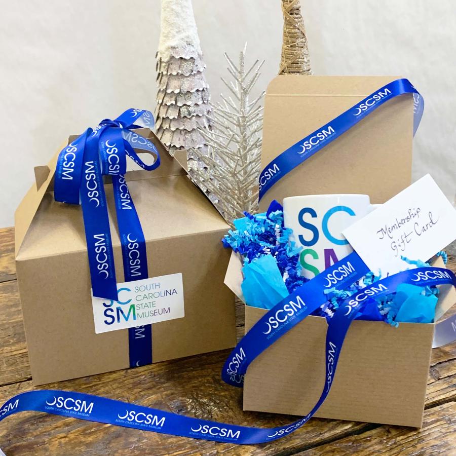 Two brown gift boxes on display with one partly open to show an SCSM mug inside with blue SCSM branded ribbon entwined
