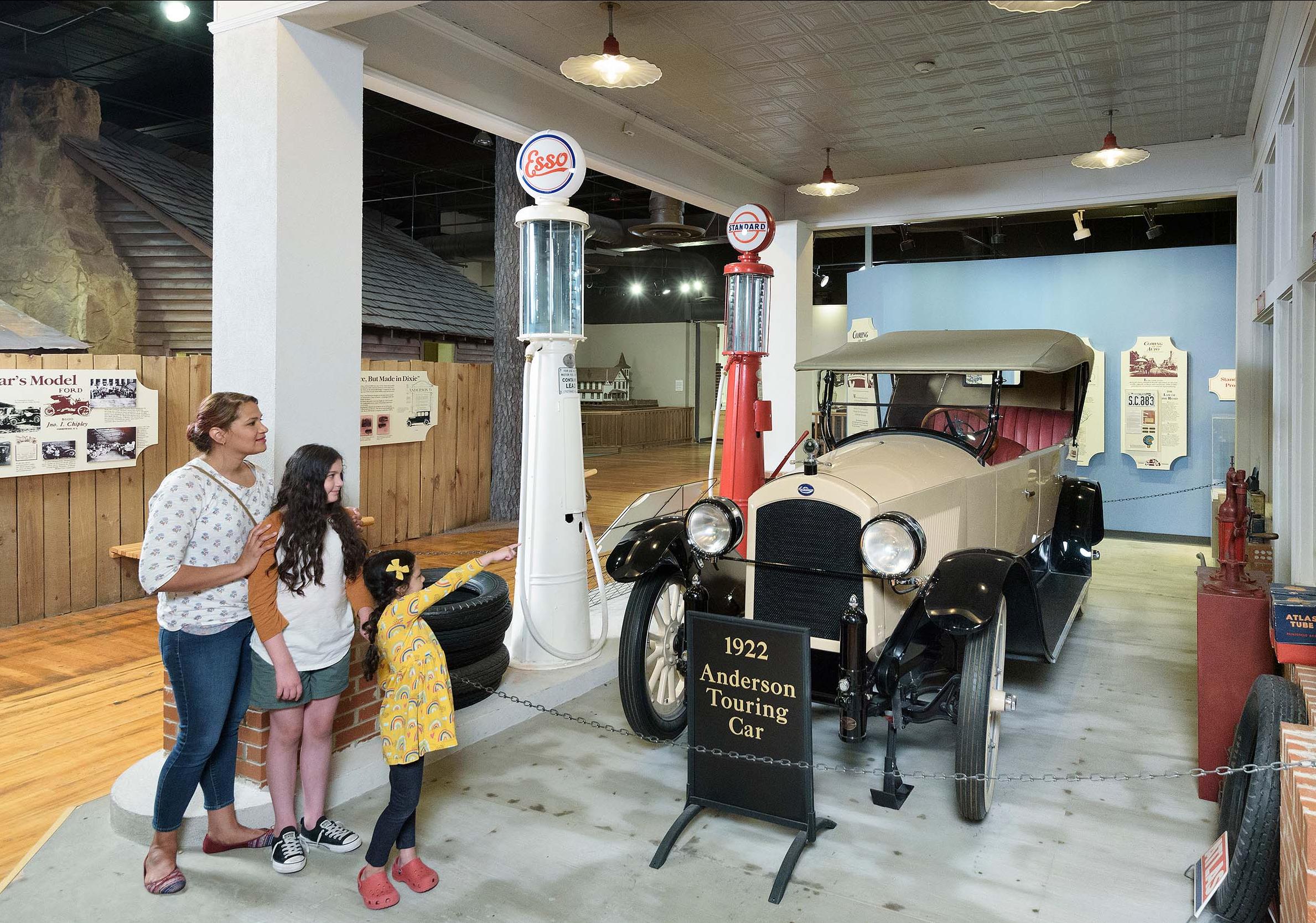 Mom and 2 daughters stand look at a white, early 20th century vintage car on display at the SC State Museum