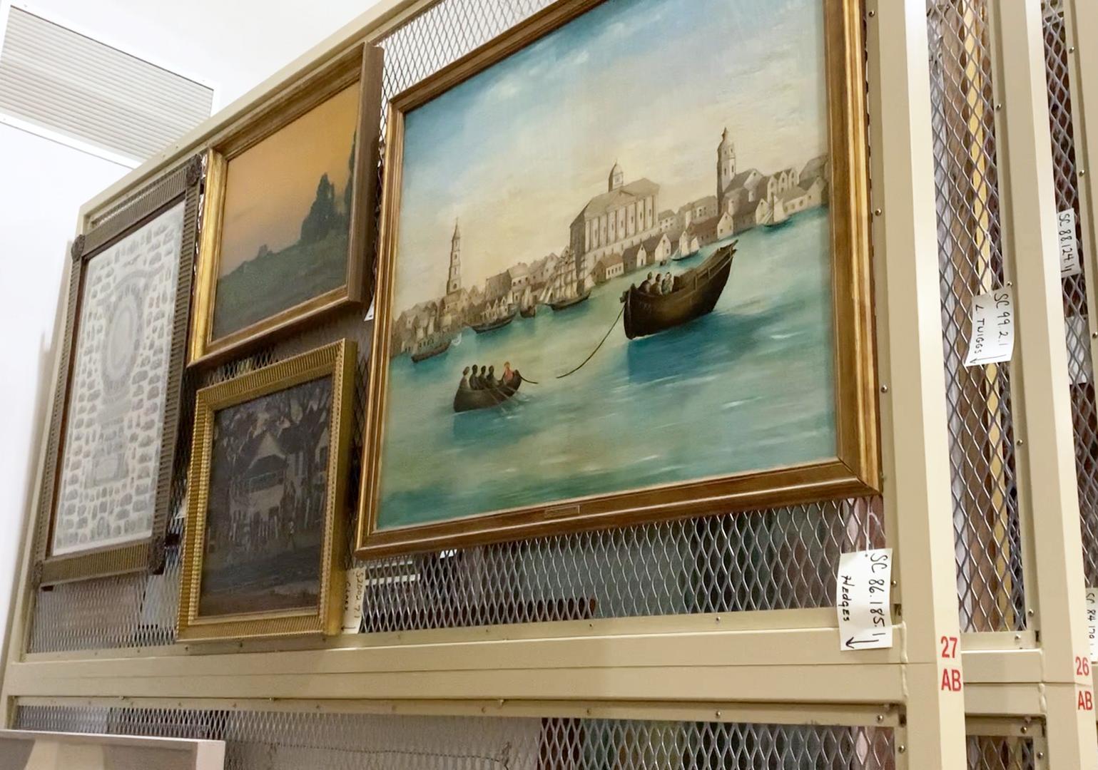 Four framed paintings are mounted on the uppermost section of a rolling rack in the museum's art storage room.