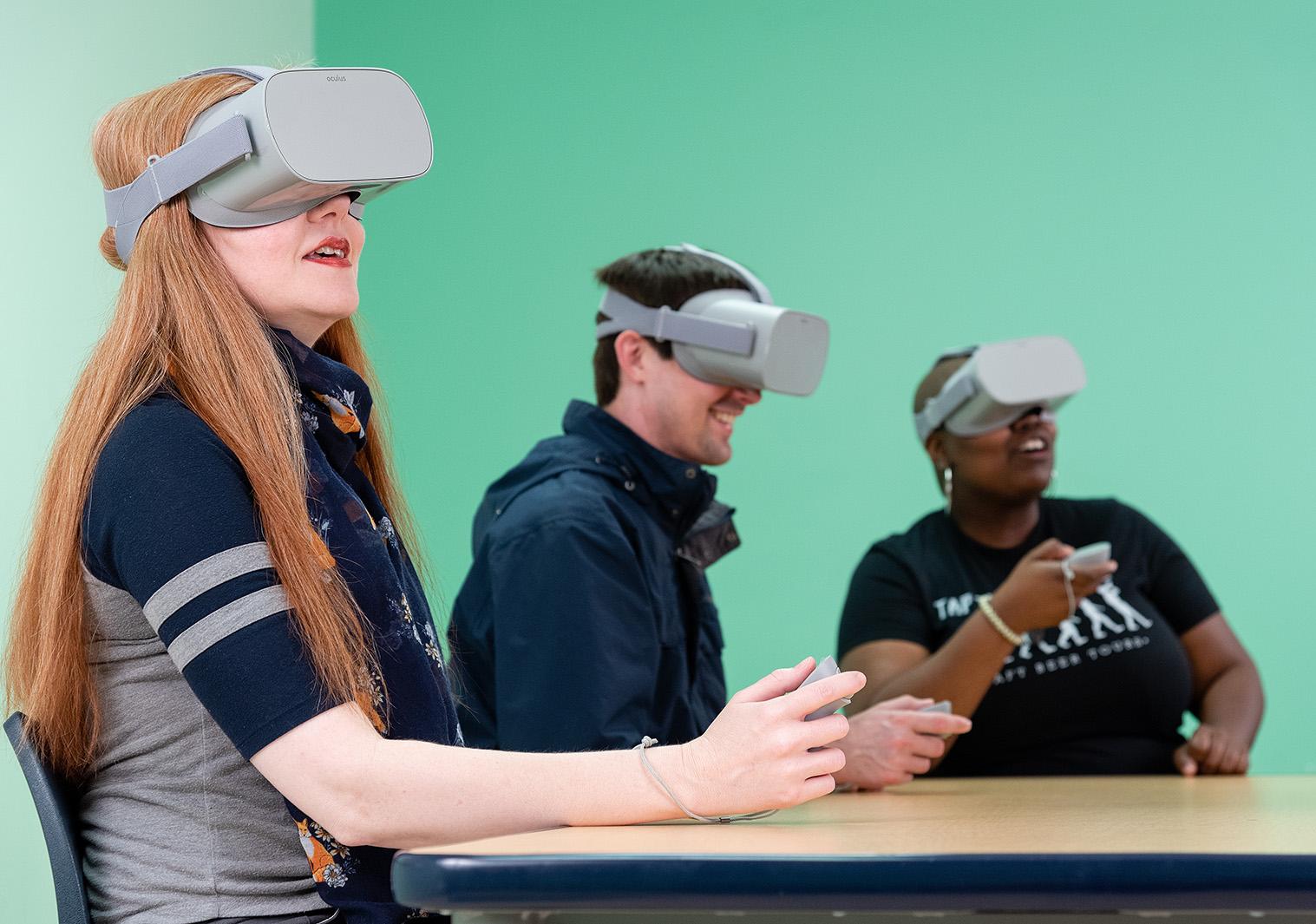 One man and two women sit a table wearing gray virtual reality head gear
