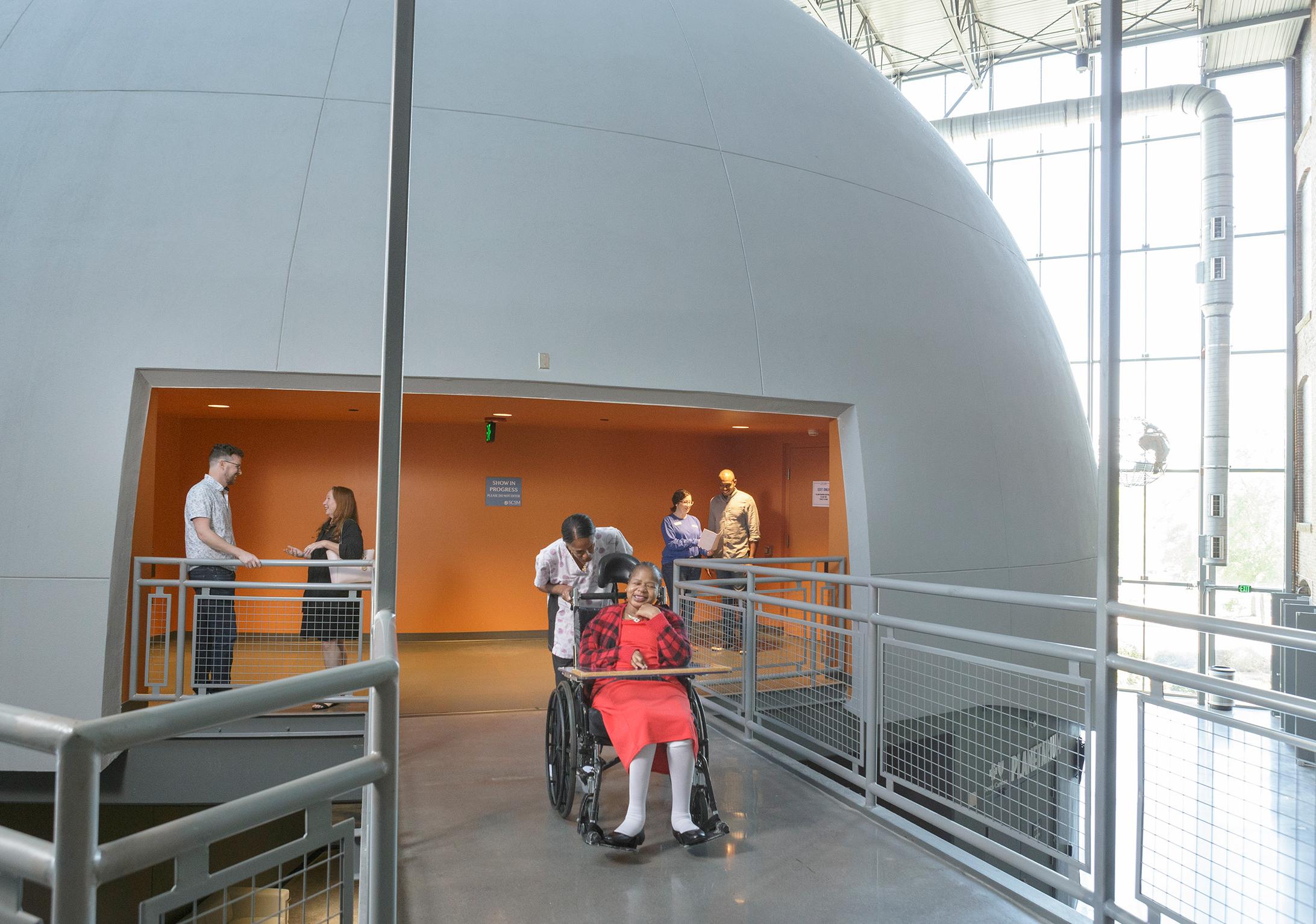 Large dome structure with exit midway up onto a catwalk space. Guest in wheelchair is on catwalk with caretaker and other guests in the background.