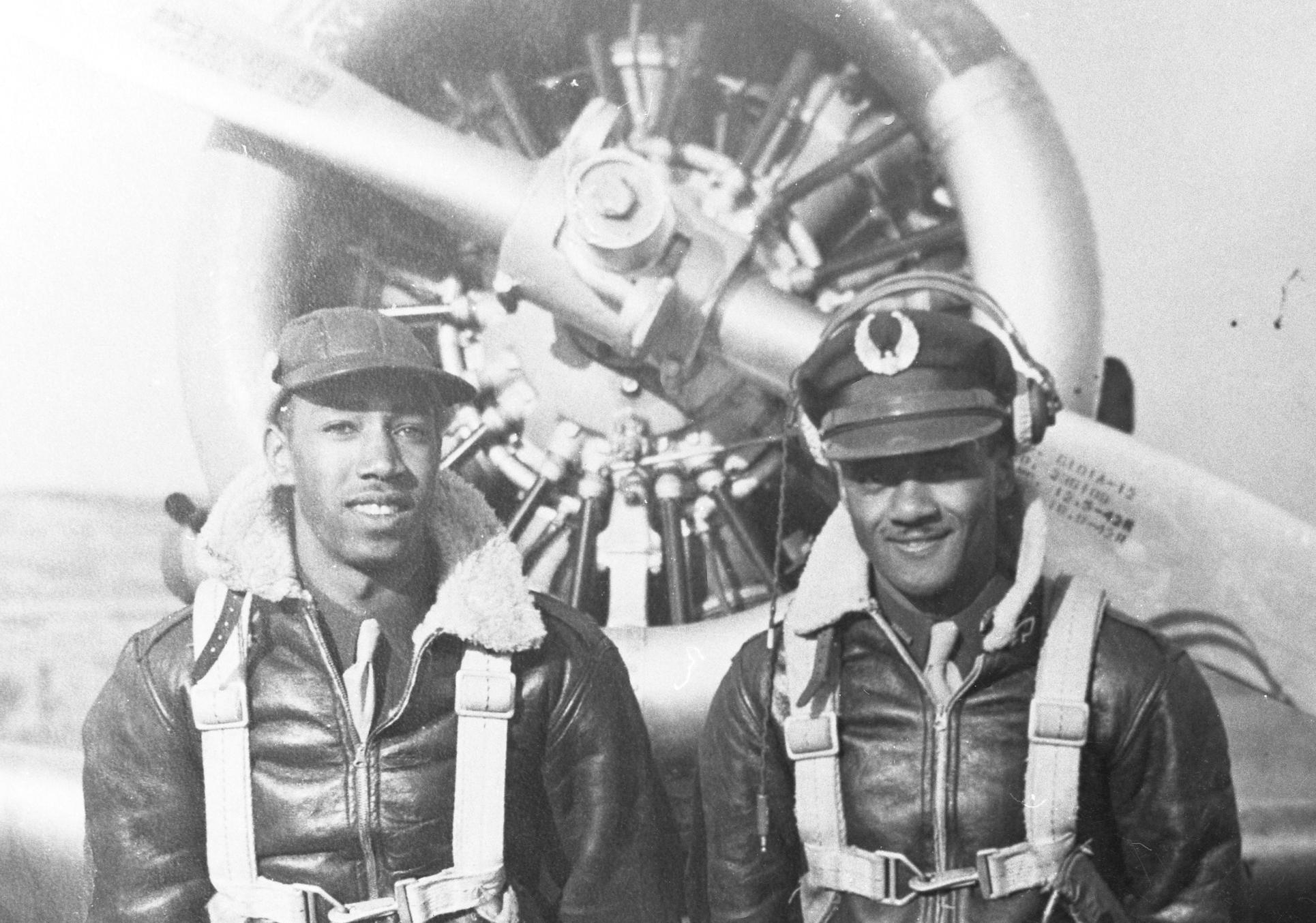 Black and white photo with two African American men wearing bomber jackets and harnesses standing in front of a propeller plane