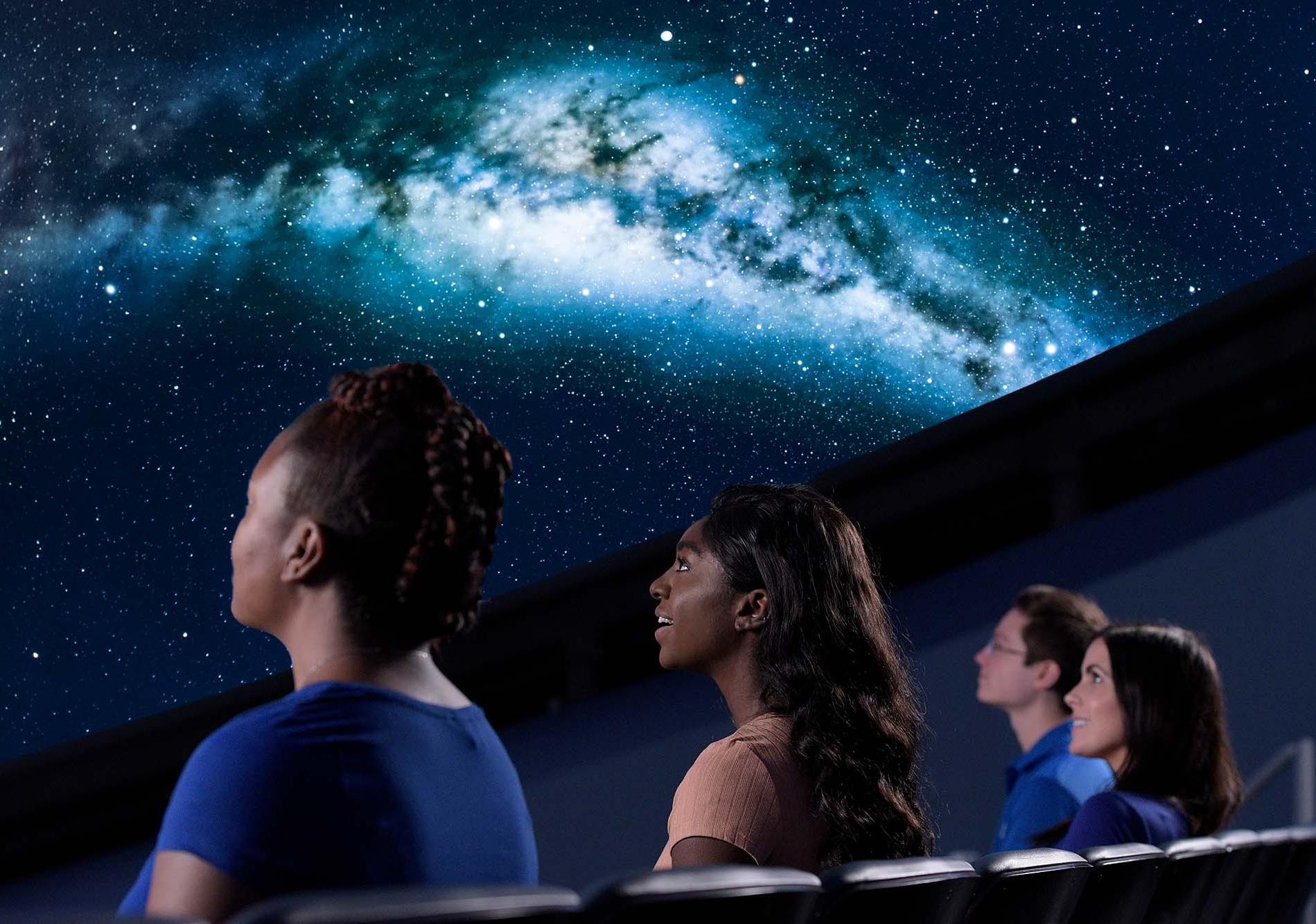 Close up image of four people shown from a back/side profile as they look up to watch a projection of the Milky Way in a planetarium dome