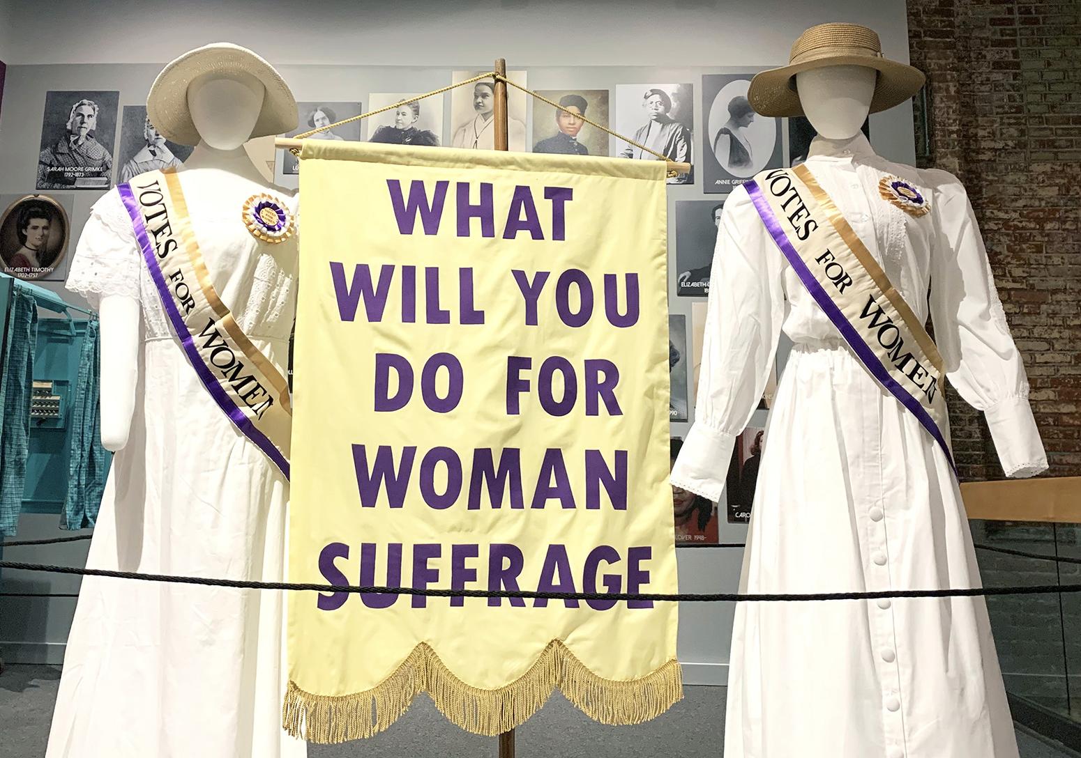 Two mannequins is white ankle length dresses wearing cream and purple trimmed sashes stand on either side of a banner that reads "What Will You Do For Woman Sufferage"