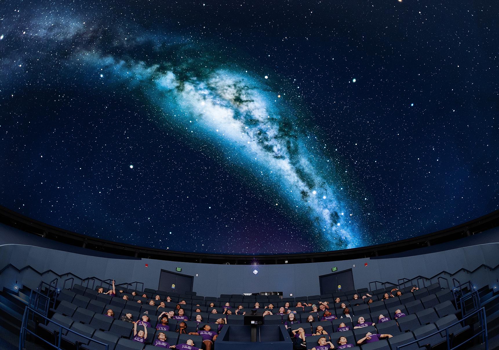 Group of school children sit under the planetarium dome which projects an image of the milky way