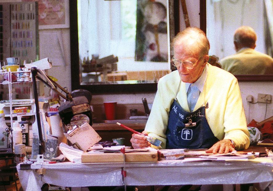 An older man with glasses with an apron over his suit coat and shirt sits at a table with a paintbrush in his hand