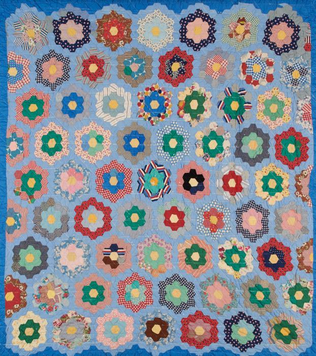 Quilt made of small hexagonal fabric pieces arranged to make flower designs in colors of blue, red, green, pink, white and yellow. Some pieces are patterned and others are solid colored.