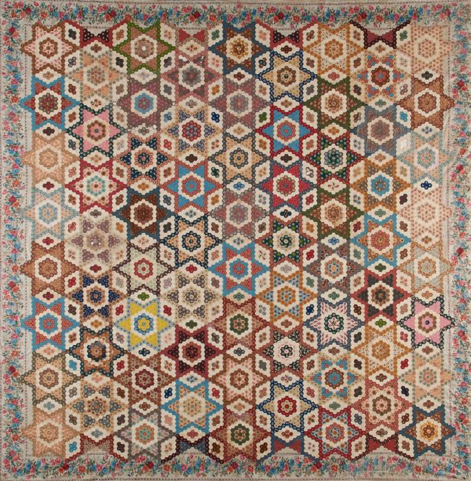Quilt made of small hexagonal fabric pieces arranged to make geometric designs in earth toned shades of blue, red, orange, yellow and brown.