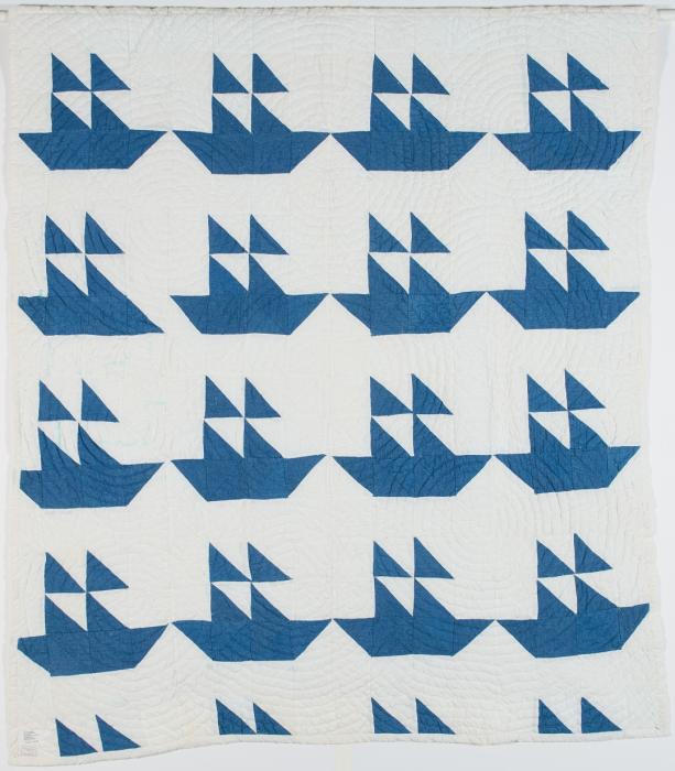 White quilt with repeated design of a ship made of triangle shaped scraps of blue fabric. There are 4 ships across and 4 down for a total of 16 ships. First ship on the second row is missing the piece of fabric that make the front bow of the ship.