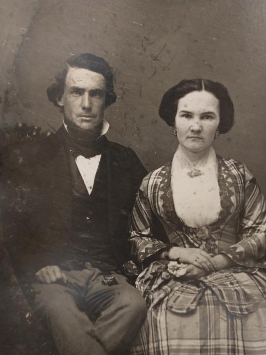Sepia toned photograph of a circa 1850 couple with man in high collar and dark colored cravat and the women in plaid dress with white blouse and matching jacket and skirt.
