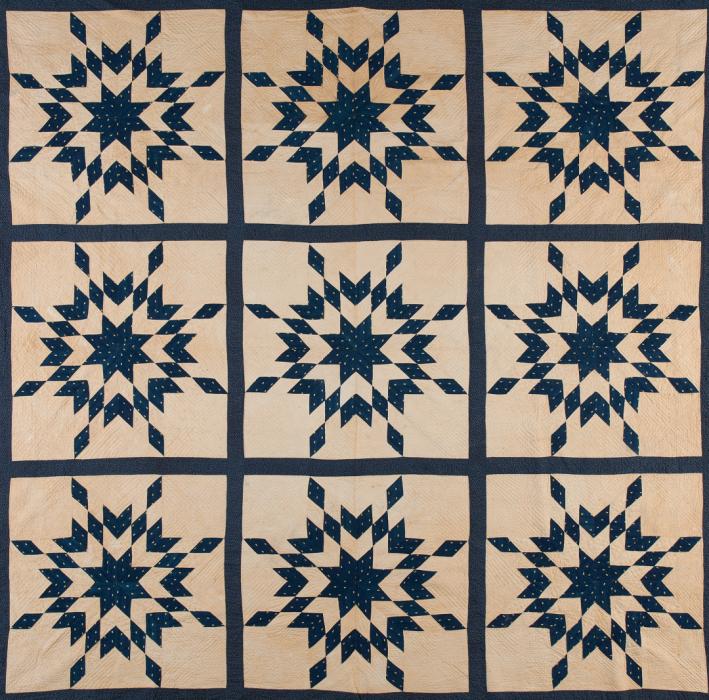 Quilt with dark blue border and cream center with dark blue lines dividing the face into 6 sections. Each section has same star shaped pattern in dark blue made out of small diamond shaped pieces.