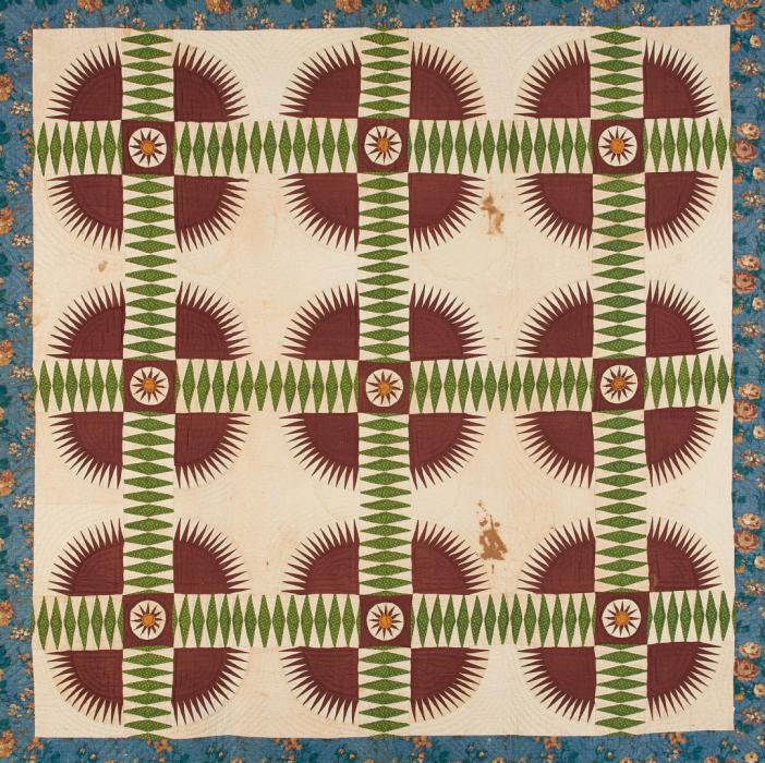 Quilt with floral pattern border and cream center with motif of red spikey circles cross cut with line made of green diamond shapes in a row.