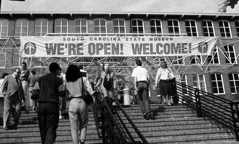 Black and white image shows guests walking up stairs towards the museum building where a banner hangs reading "We're Open! Welcome!"