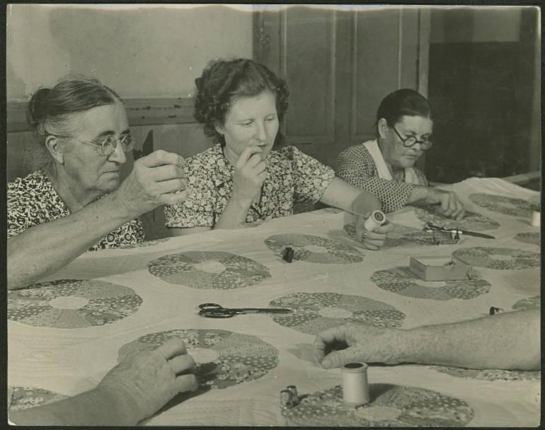 Black and white image of three women sitting at a table sewing a quilt together.