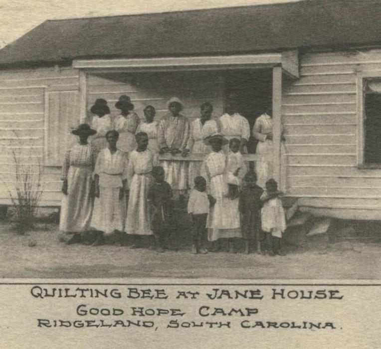 Sepia toned image shows group of women in light colored dresses on the porch of a wooden house. Underneath the image it read "Quilting Bee at Jane House, Good Hope Camp, Ridgeland, SC."